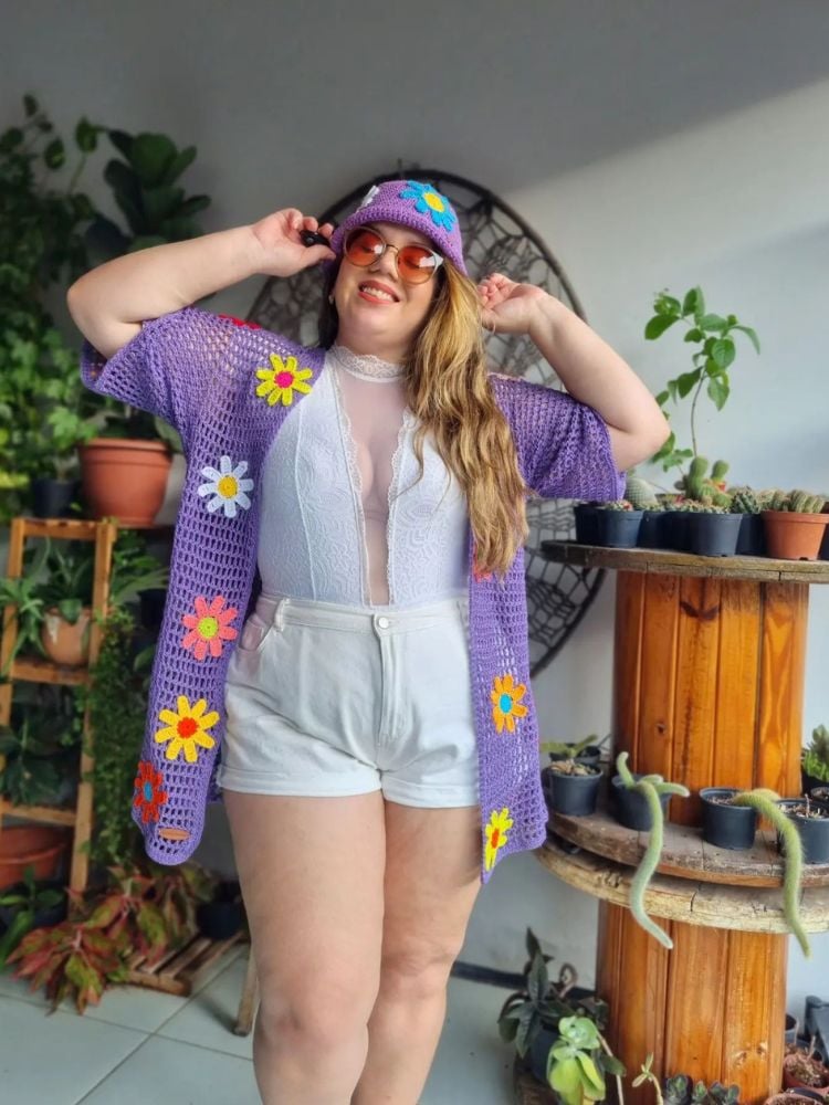 Mulher de pele clara usando body branco com renda, shorts branco, cardigan lilás de crochê com flores e bucket hat lilás de crochê com flores