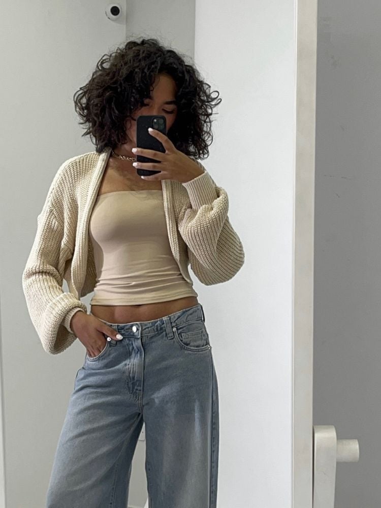 Mulher de pele morena usando cropped faixa largo bege, casaco cropped de tricô e calça jeans