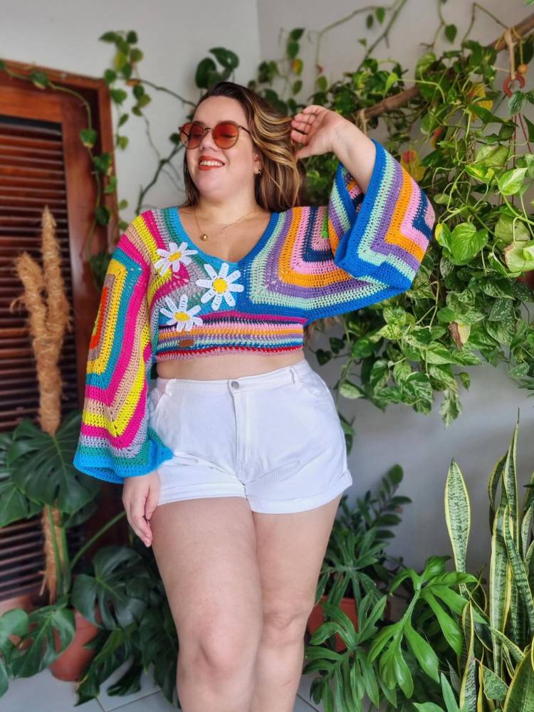 Mulher de pele clara usando blusa de manga comprida colorida com margaridas, tudo em crochê, e shorts branco