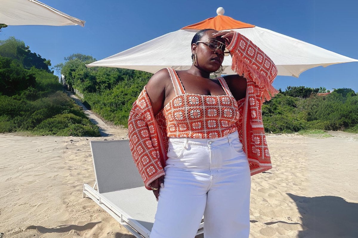 Mulher de pele negra na praia usando calça branca, top rosa de crochê e cardigan de crochê igual ao cropped