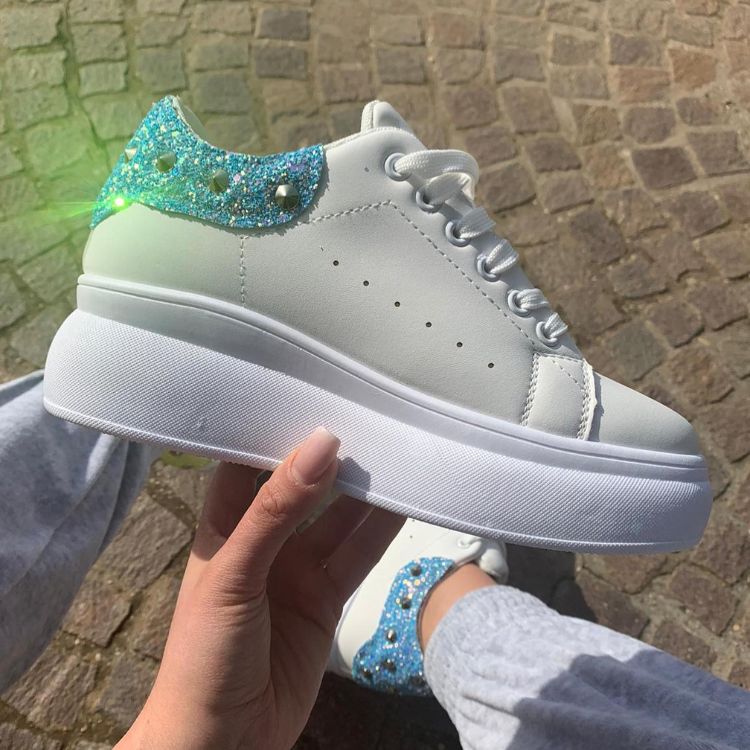 Pessoa de pele clara segurando tênis branco com parte de trás com glitter azul e spikes