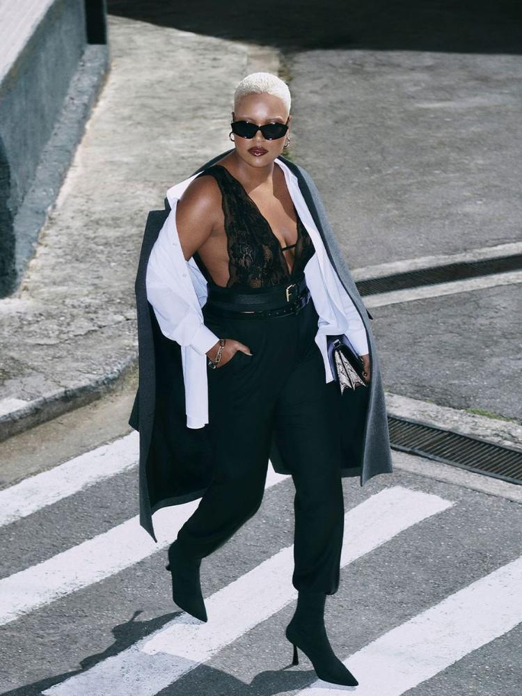 Mulher de pele negra com body preto transparente, camisa branca, sobretudo preto e calça preta