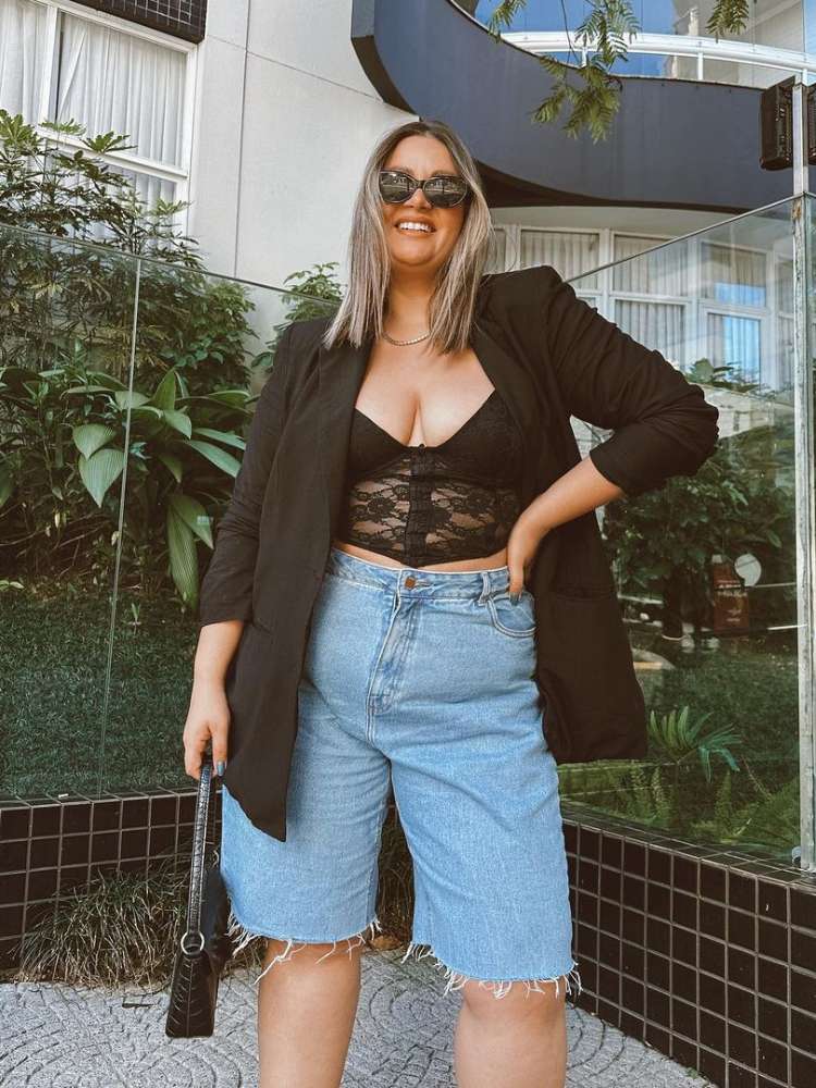 Mulher de pele morena usando corset preto de renda com transparência, blazer preto e bermuda jeans