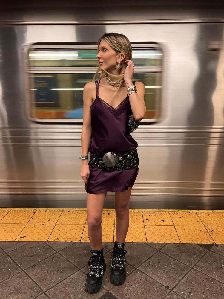 Mulher de pele clara na plataforma do metrô usando camisola vinho, cinto grande preto e acessórios