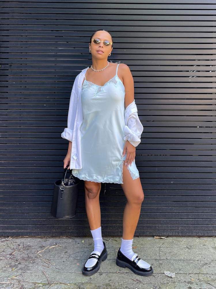 Mulher de pele morena usando camisola branca e robe branco, com sapato preto e meias brancas cano médio