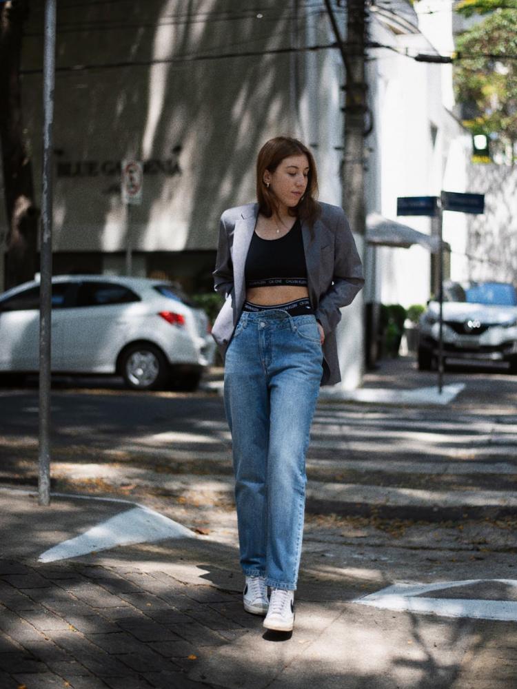 Mulher de pele clara usando blazer cinza aberto, top preto e calça jeans com cós de calcinha à mostra