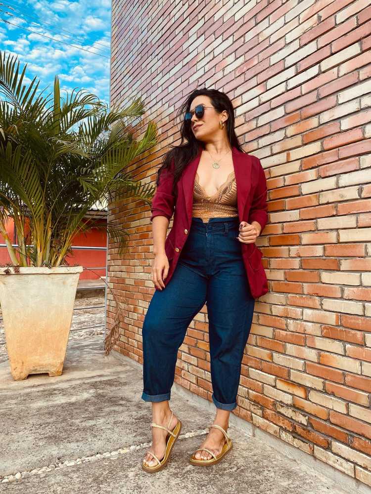 Mulher de pele clara usando sutiã dourado, blazer vinho e calça jeans justa