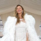 A imagem mostra Gisele Bündchen sorridente usando um elegante vestido de festa branco, adornado com penas. O ambiente é sofisticado, com lustres de cristal ao fundo. O vestido é justo, destacando a silhueta, e o casaco de penas adiciona um toque glamoroso ao visual.