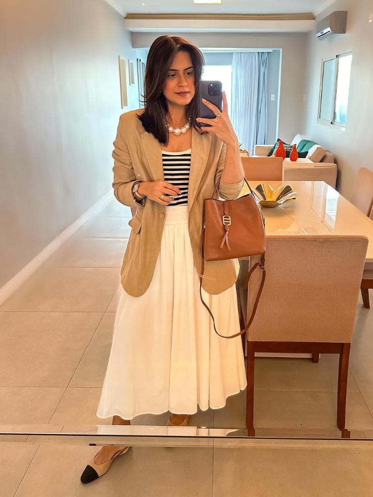 Mulher de pele clara tirando foto no espelho usando blazer bege, blusa listrada branca e preta e saia midi branca 