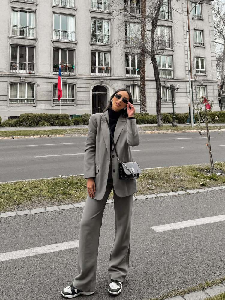 Mulher de pele clara usando blusa preta gola alta, blazer e calça cinzas e tênis 