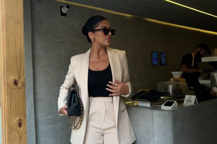 Mulher de pele clara em cafeteria usando blazer e calça social beges e blusa preta, além de bolsa preta e óculos de sol