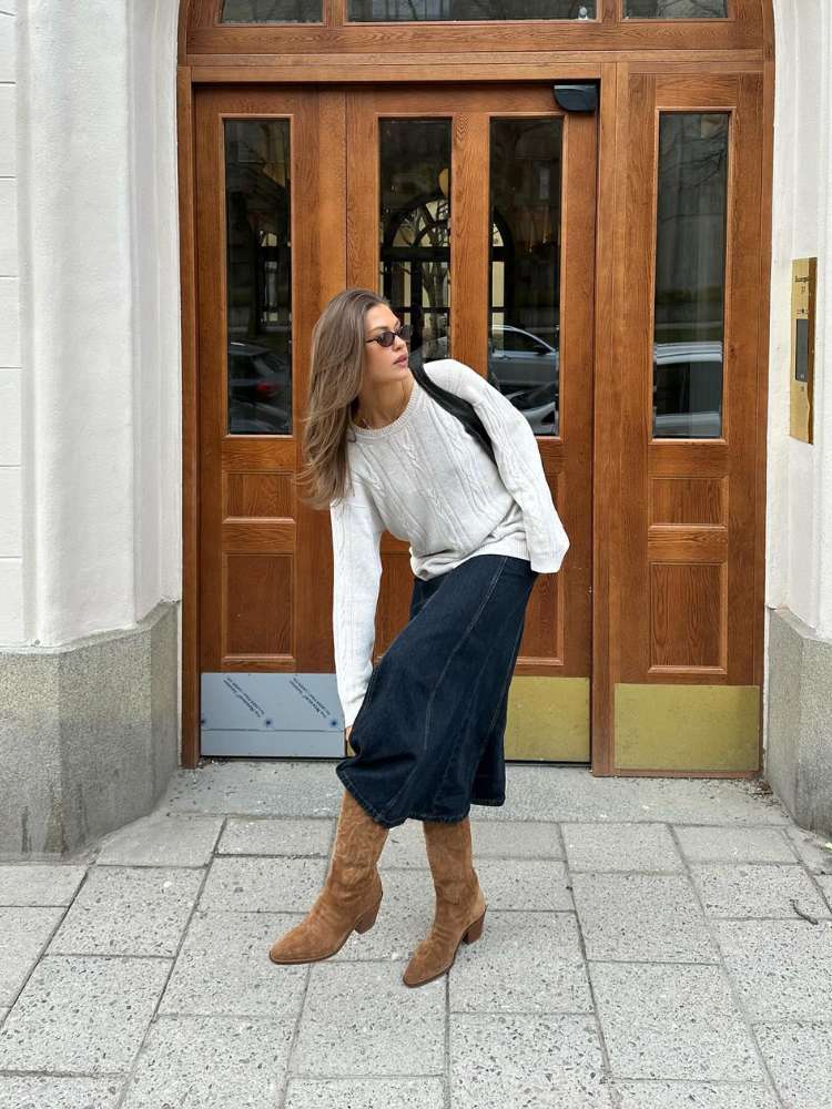 Mulher de pele clara usando suéter off white, saia midi jeans e bota western marrom 