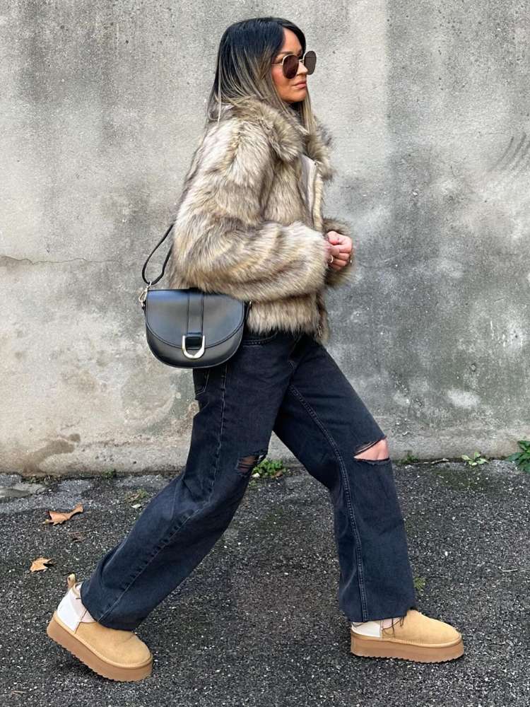 Mulher de pele clara usando casaco de pele bege, calça jeans escura, bolsa de couro preta e ugg boots 