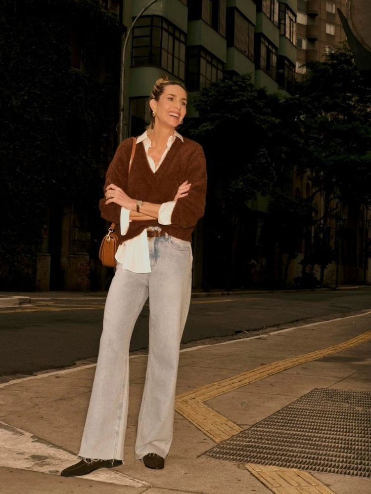 Mulher de pele clara usando calça jeans clara, camisa branca por cima de suéter marrom e bolsa marrom