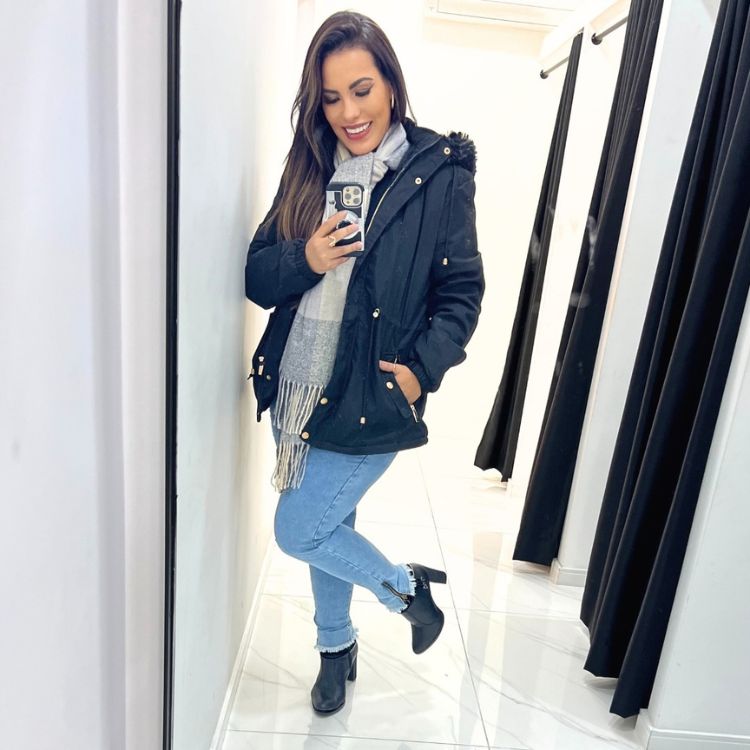 Mulher de pele clara tirando foto no espelho usando parka preta, calça jeans, cachecol cinza e bota preta 