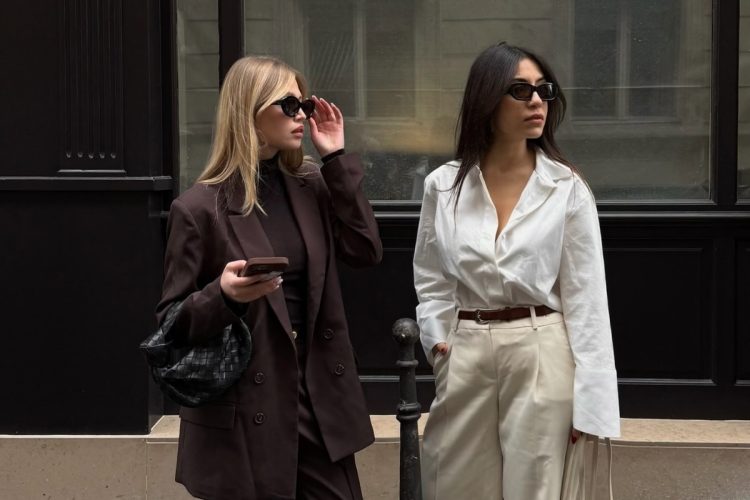 Duas mulheres posam elegantemente na rua. A primeira usa terno marrom, segura um celular e bolsa preta. A segunda veste camisa branca e calça bege. Ambas usam óculos de sol. Fundo com muro preto e vidro.