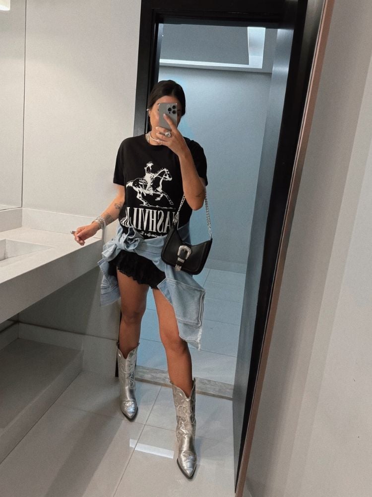 Mulher de pele clara tirando foto no espelho com camiseta preta, saia preta, bota branca e jaqueta amarrada na cintura 