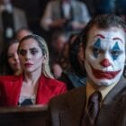 Cena de "Coringa: Delírio a Dois" em que Lady Gaga de Harley Quinn está sentada atrás de Joaquim Phoenix de Coringa. Ela usa roupa vermelha e maquiagem preta e ele usa terno cinza e cabelo verde, rosto branco, sobrancelha vermelha, nariz vermelho, boca vermelha, sombra azul