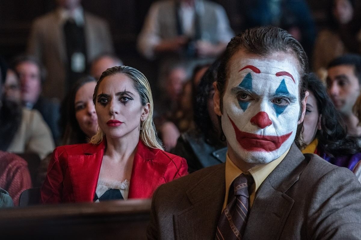 Cena de "Coringa: Delírio a Dois" em que Lady Gaga de Harley Quinn está sentada atrás de Joaquim Phoenix de Coringa. Ela usa roupa vermelha e maquiagem preta e ele usa terno cinza e cabelo verde, rosto branco, sobrancelha vermelha, nariz vermelho, boca vermelha, sombra azul