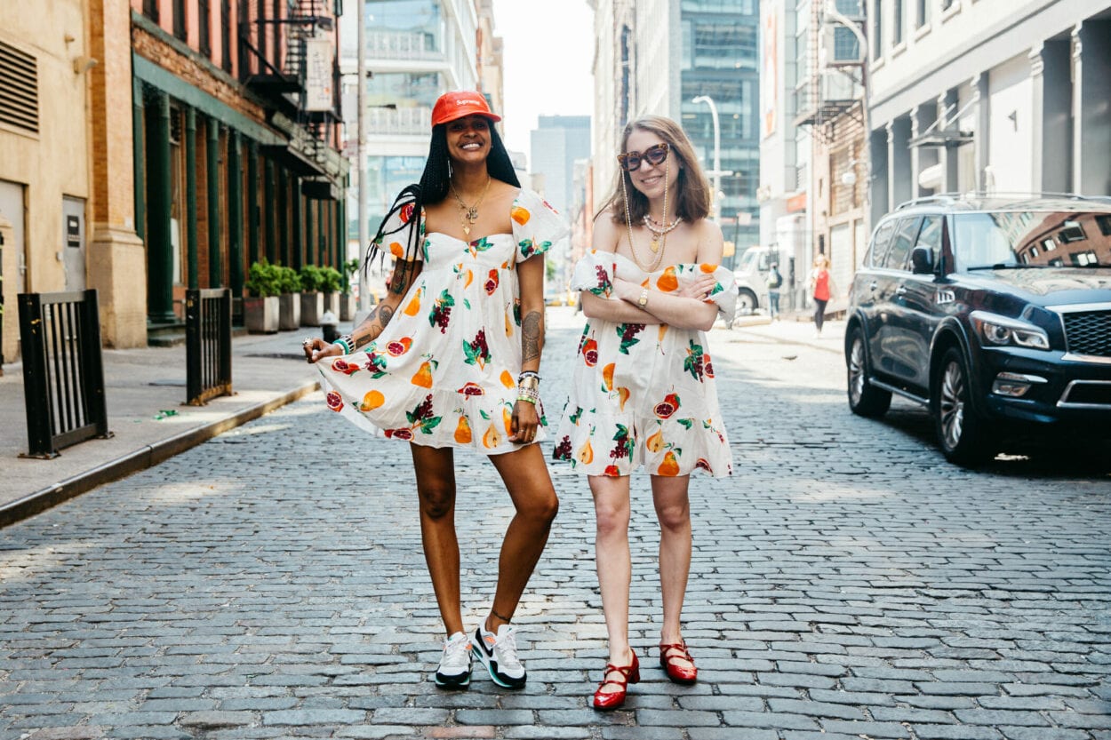 Duas mulheres, uma de pele negra e outra de pele clara, usando vestidos beges com estampa de frutas 