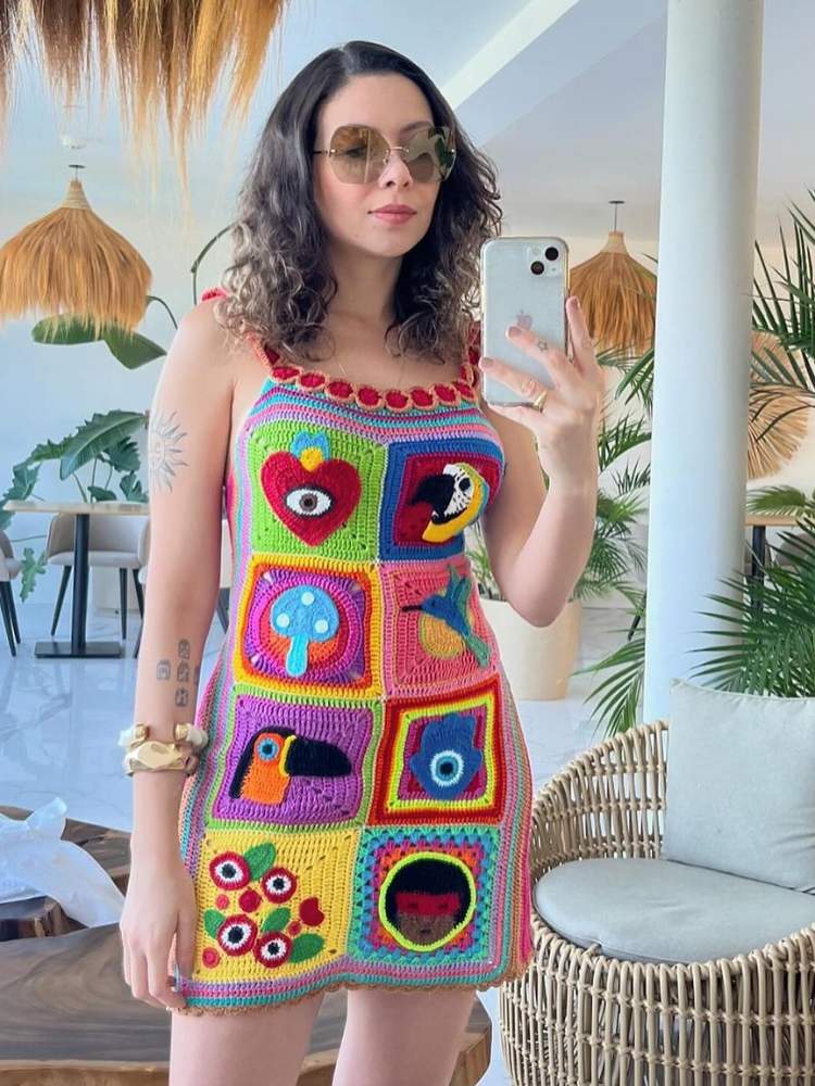 A imagem mostra uma mulher de pele clara tirando uma selfie em um ambiente moderno. Ela está vestindo um vestido colorido feito de diferentes patches de crochê, cada um com designs únicos, como olhos, flores e padrões abstratos. O ambiente tem uma decoração moderna com móveis de vime e luminárias penduradas que se assemelham a cestos invertidos.