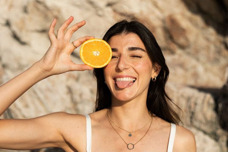 Mulher de pele clara em frente à pedras, segurando metade de uma laranja e sorrindo com a língua para fora