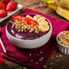 Foto de tigela de açaí com granola e morango sob toalha de mesa vinho e ingredientes da tigela ao redor