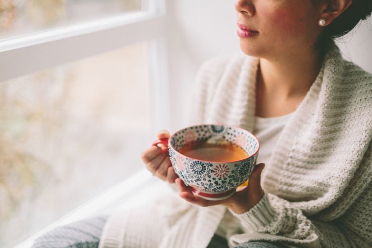 Mulher de pele clara sentada próxima à janela, usando casaco bege e segurando xícara colorida com chá
