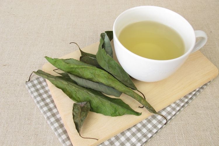 Mesa clara com toalha cinza xadres, tábua de madeira, xícara branca com chá verde e folha de abacate