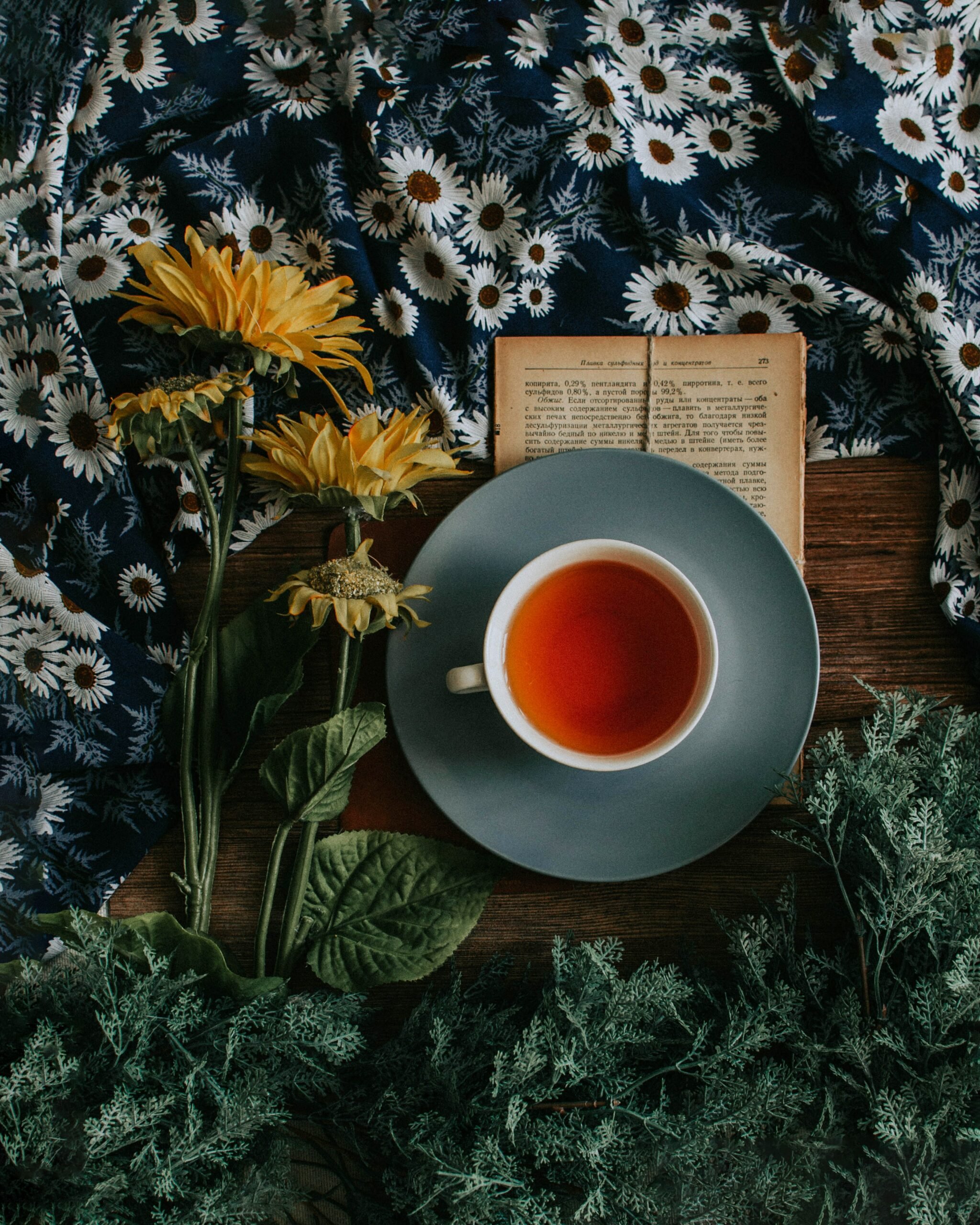 Foto de toalha de margaridas, girassóis, livro aberto, chá laranja em xícara e pires azul e ervas