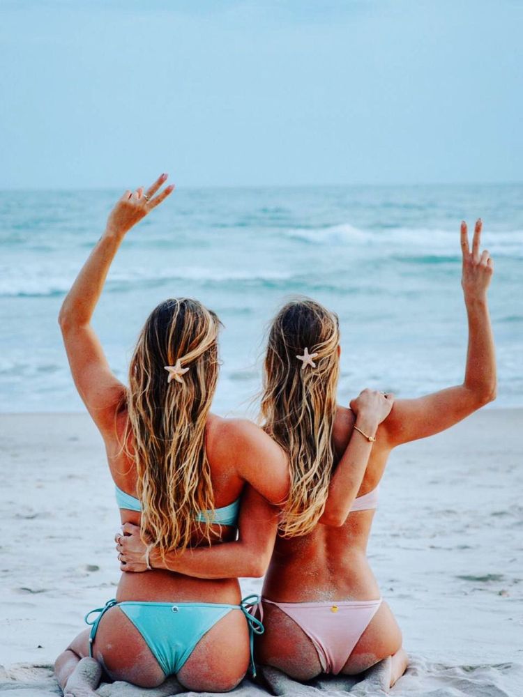 Duas mulheres loiras de pele clara de costas para a foto e de frente para o mar, usando concha no cabelo