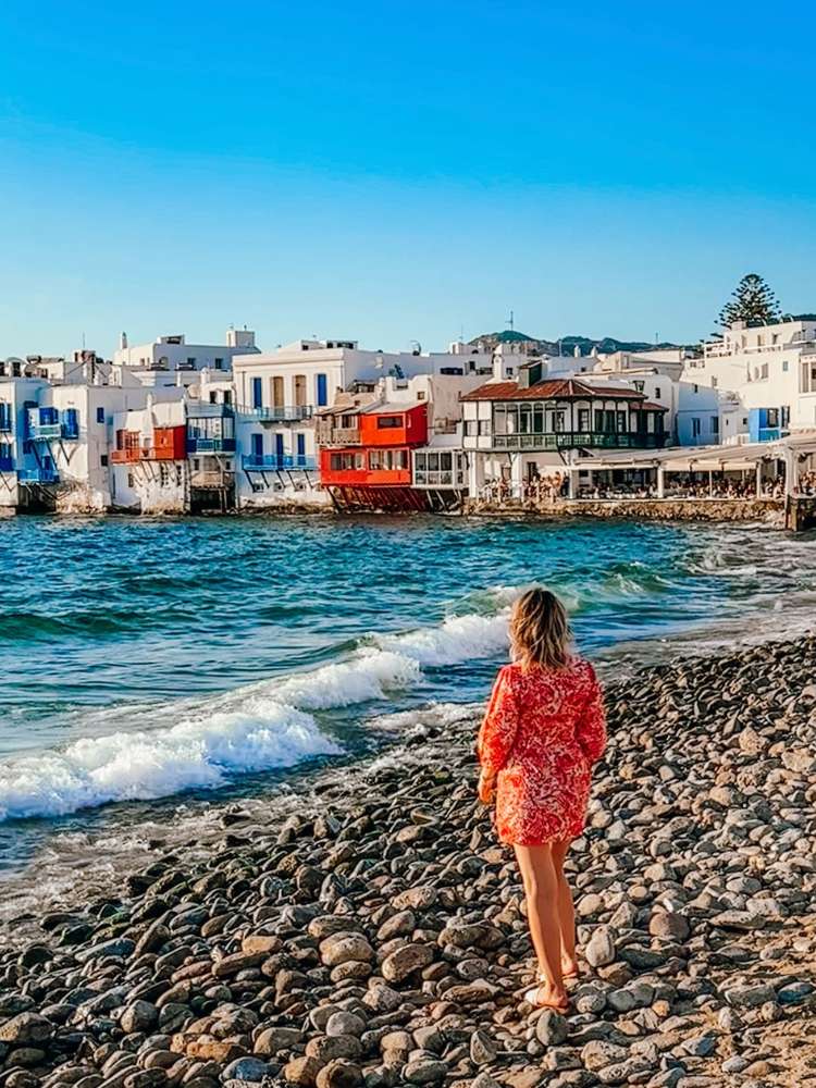 Mulher loira de pele clara usando vestido laranja nas pedras de praia na Grécia, com casas coloridas ao fundo