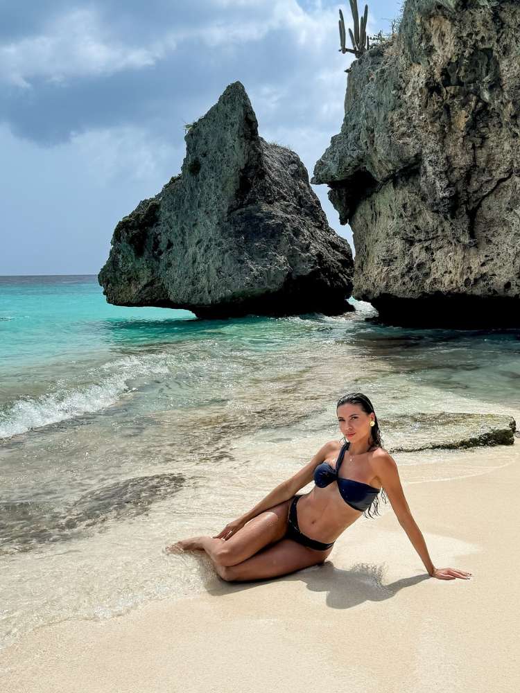 Mulher de pele clara usando biquini preto, deitada na areia à beira do mar com pedras ao fundo