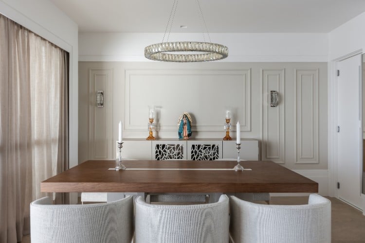 Sala de jantar com mesa de madeira, poltronas cinzas, lustre, parede branca e bege com boiserie