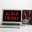 Fundo cinza com notebook escrito black friday de vermelho em imagem preta. Bolsa preta e mini carrinho de compras