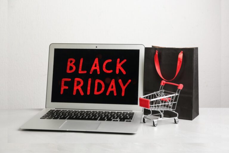 Fundo cinza com notebook escrito black friday de vermelho em imagem preta. Bolsa preta e mini carrinho de compras