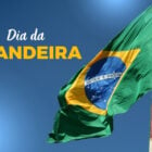 A imagem mostra a bandeira do Brasil tremulando ao vento, destacando suas cores verde, amarelo, azul e branco. No centro, o lema "Ordem e Progresso" é visível. O fundo é um céu azul claro. À esquerda, está escrito "Dia da Bandeira" em letras brancas e amarelas.