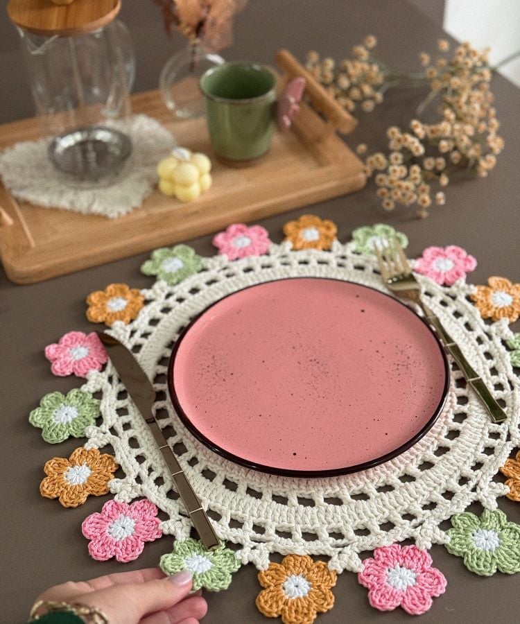 Um sousplat de crochê branco com bordas decoradas com pequenas flores coloridas em rosa, verde e laranja. Sobre ele, há um prato rosa, com talheres dourados ao lado. Ao fundo, uma bandeja de madeira com um bule de vidro, uma xícara verde e flores secas.