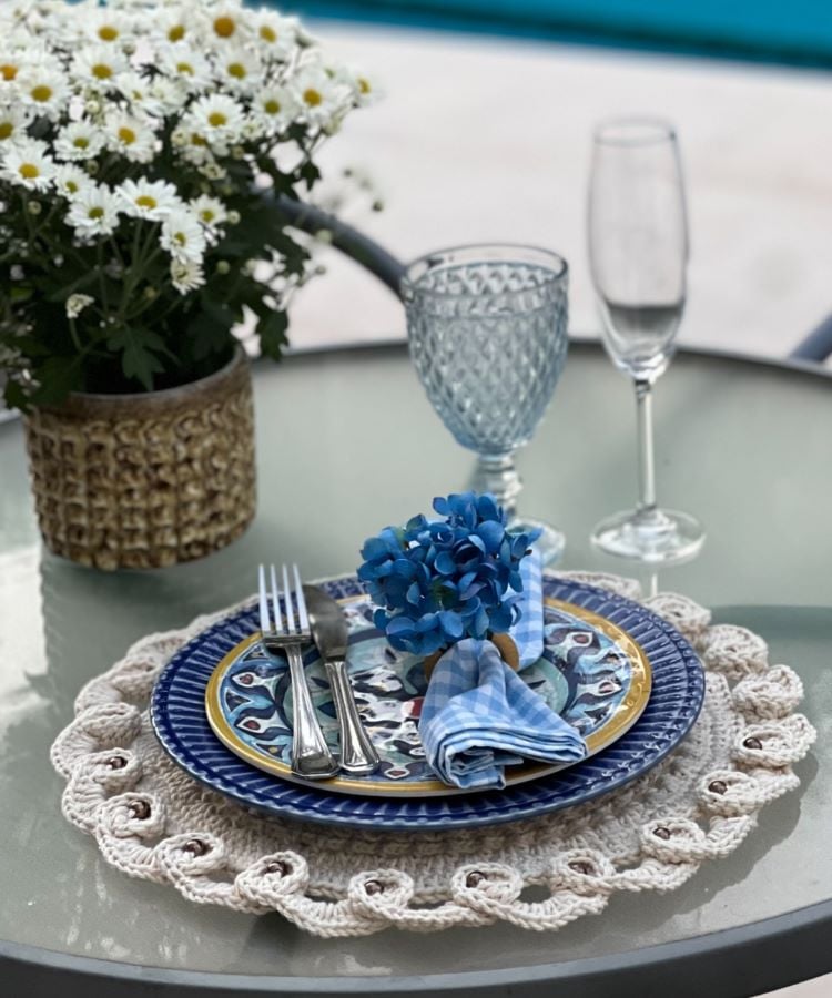 Uma mesa redonda de vidro com um sousplat de crochê branco, decorado com um prato azul, talheres e um guardanapo azul com flores. Ao lado, há um vaso de margaridas brancas, uma taça de cristal azul e uma taça de champanhe.