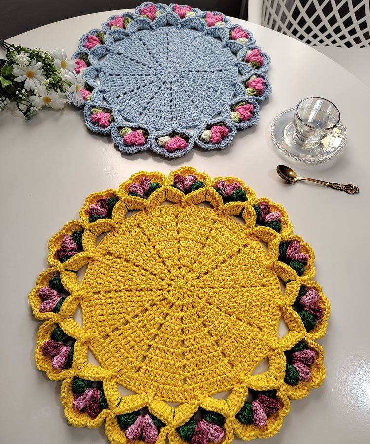 Dois sousplats de crochê em uma mesa branca. Um é amarelo com bordas de flores rosa e verde, e o outro é azul com bordas de flores rosa e verde. Ao lado, há uma xícara de chá transparente e uma colher dourada.