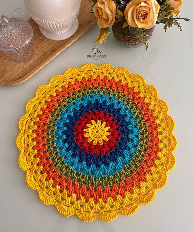 Um sousplat de crochê colorido com tons de amarelo, laranja, vermelho, azul e verde, em formato circular e com bordas onduladas. Está sobre uma mesa de vidro, ao lado de um vaso com rosas amarelas e um pote de vidro transparente.