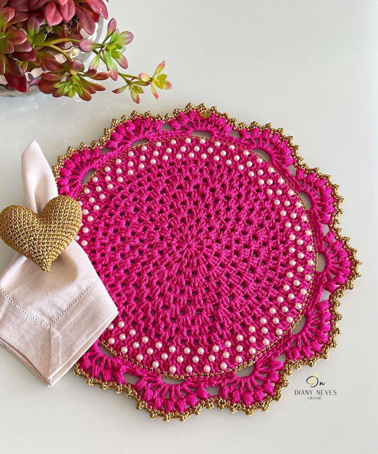 Um sousplat de crochê rosa-choque com bordas douradas e detalhes em pérolas brancas. Está sobre uma mesa de vidro, acompanhado de um guardanapo branco com um porta-guardanapo dourado em formato de coração, e um vaso com plantas suculentas.