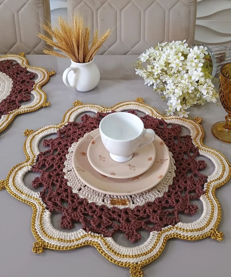 Um sousplat de crochê com bordas onduladas em marrom, bege e dourado. Sobre ele, há um prato bege decorado com folhas marrons e uma xícara branca. Ao fundo, um vaso branco com trigo seco e outro com flores brancas. Uma taça âmbar completa a decoração.