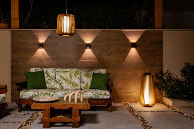 Espaço zen ao ar livre, com muro marrom, opções de iluminação, sofá de fundo bege com folhas estampadas, almofadas verdes, mesa de centro de madeira e vaso com plantas