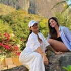 Duas mulheres posam sorridentes em um ambiente natural. Uma veste branco com boné, a outra uma camisa listrada sobre roupa preta. Estão sentadas em uma pedra, cercadas por vegetação exuberante com flores vermelhas e palmeiras, ao fundo uma montanha.