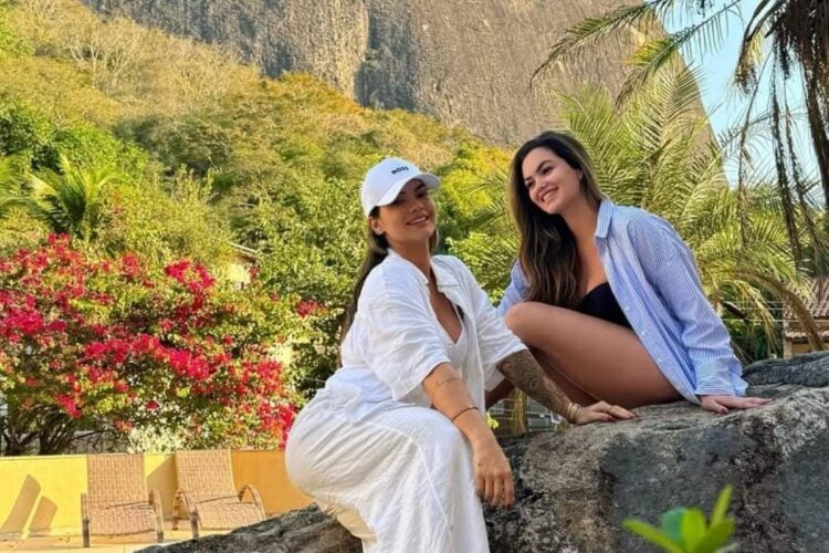 Duas mulheres posam sorridentes em um ambiente natural. Uma veste branco com boné, a outra uma camisa listrada sobre roupa preta. Estão sentadas em uma pedra, cercadas por vegetação exuberante com flores vermelhas e palmeiras, ao fundo uma montanha.