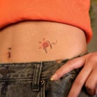 A imagem mostra a parte inferior do abdômen de uma pessoa, com um top laranja e jeans. Há um piercing no umbigo e uma tatuagem delicada de flor com traços finos e pétalas vermelhas. A mão está levemente posicionada sobre o cós da calça, destacando o estilo casual e jovem.