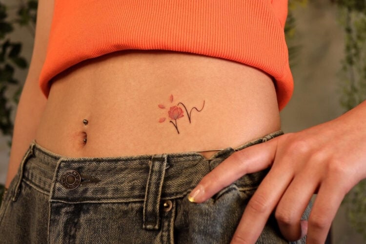 A imagem mostra a parte inferior do abdômen de uma pessoa, com um top laranja e jeans. Há um piercing no umbigo e uma tatuagem delicada de flor com traços finos e pétalas vermelhas. A mão está levemente posicionada sobre o cós da calça, destacando o estilo casual e jovem.