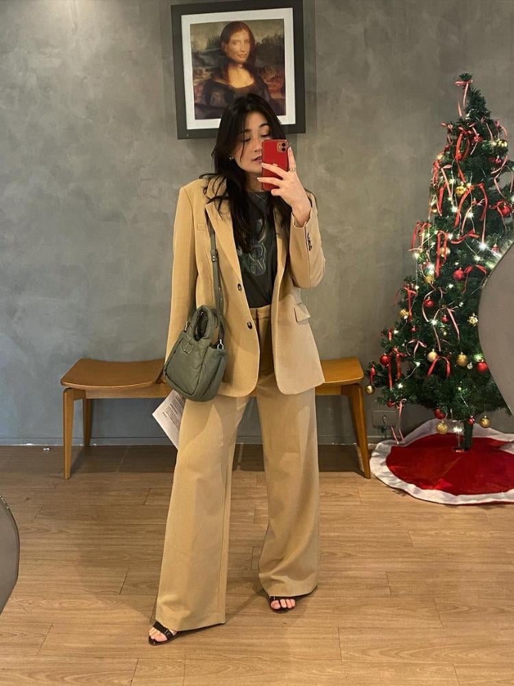 Mulher de descendência asiática em local com parede cinza e árvore de natal, usando blusa cinza, bolsa cinza e blazer com pantalona, ambos beges