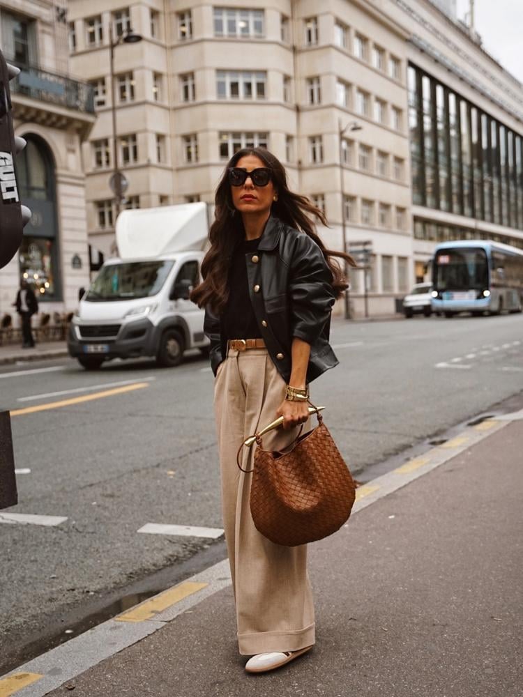 Mulher de pele clara na calçada usando pantalona bege, blusa preta, jaqueta preta de couro, bolsa marrom e acessórios